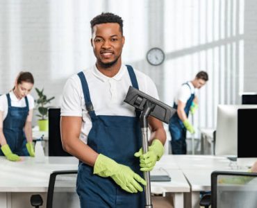 smiling african american cleaner holding vacuum cleaner brush near team of multicultural colleagues