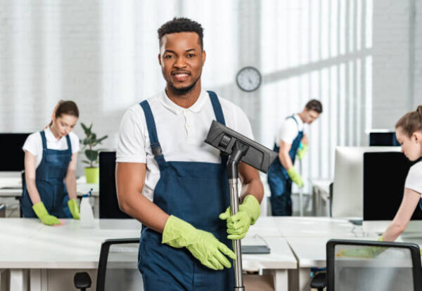 smiling african american cleaner holding vacuum cleaner brush near team of multicultural colleagues