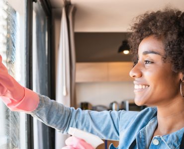 Portrait of young afro woman in gloves cleaning window with rag at home. Housework, housekeeping and cleaning concept.